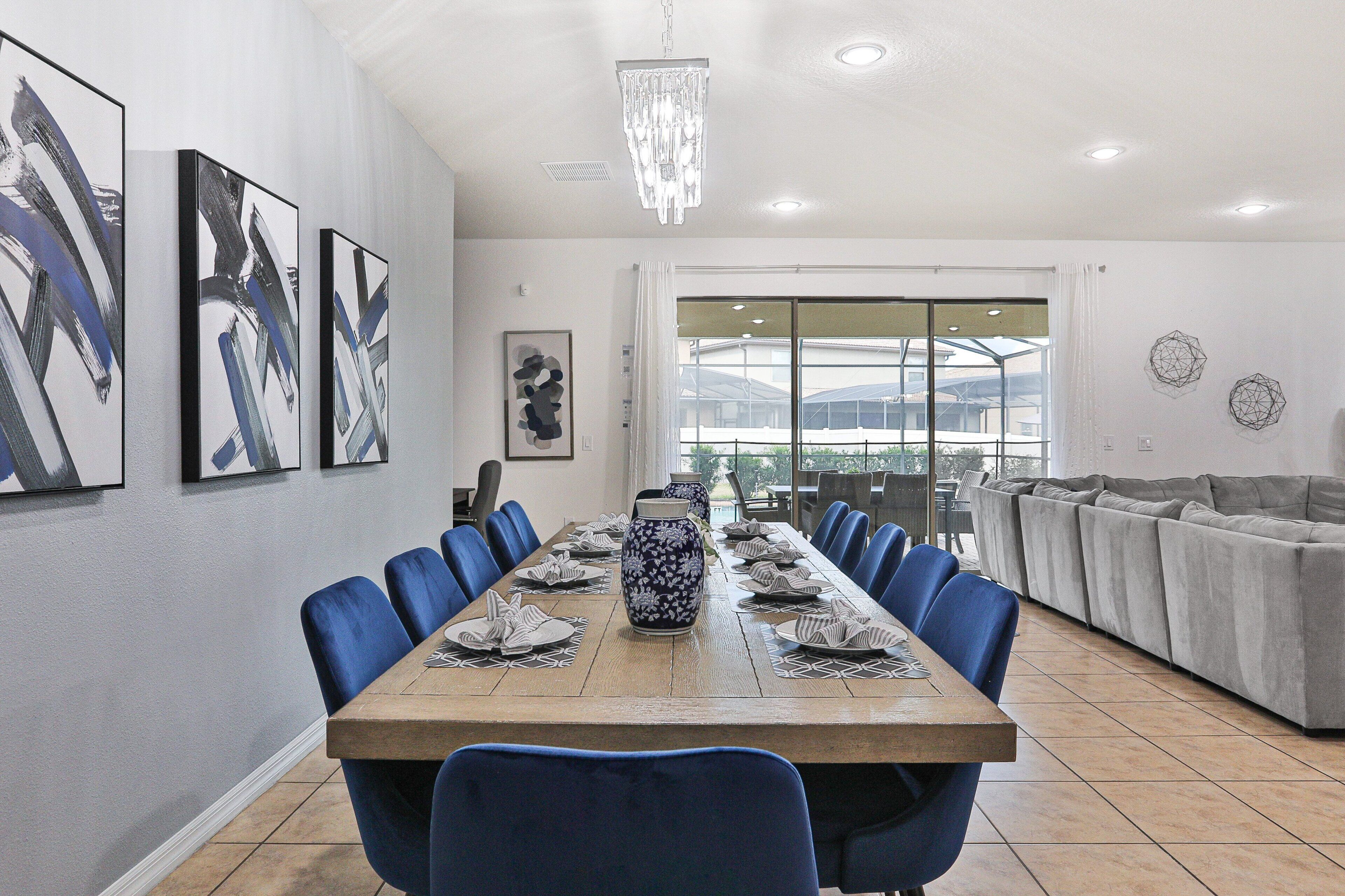 A dining area with a long table set for six, blue chairs, a vase centerpiece, and a chandelier.