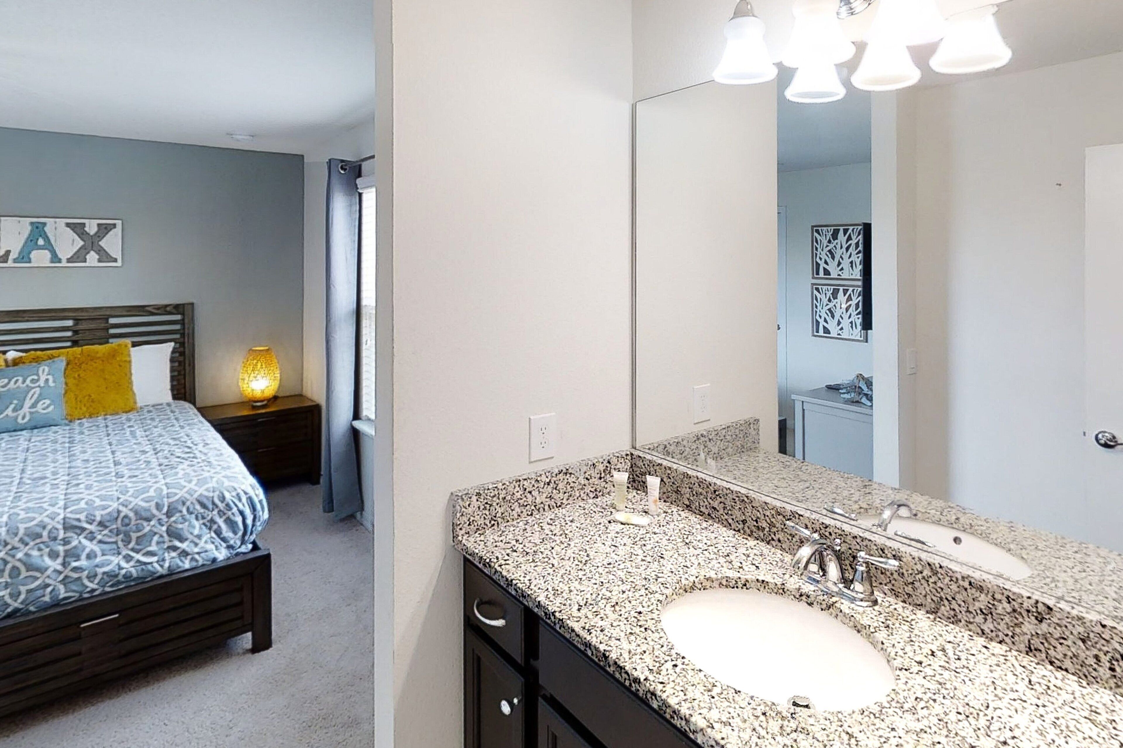 A bedroom with a bed, bedside table, lamp, and a large mirror above a granite countertop sink.