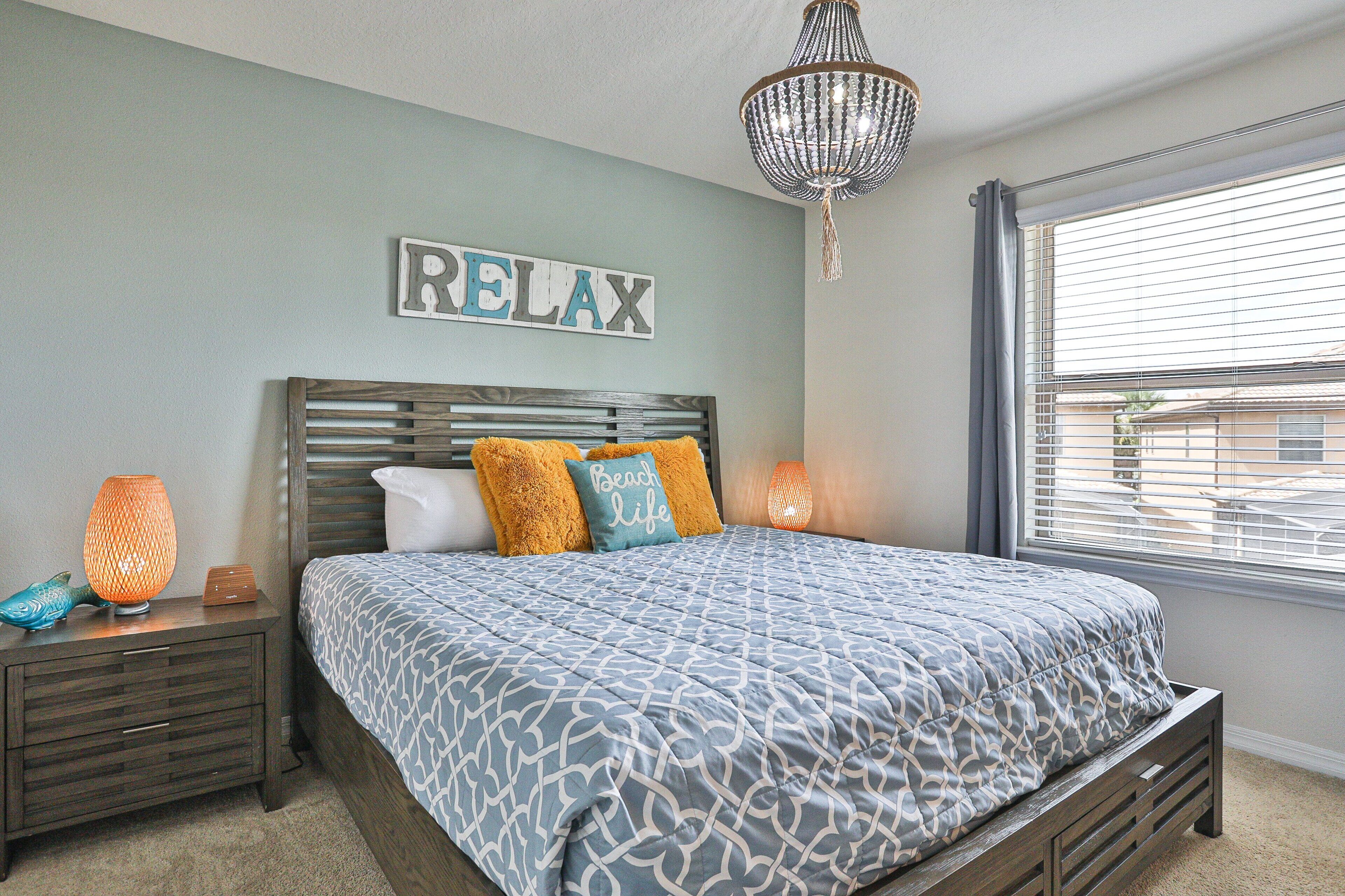 A bedroom with a bed, a nightstand, a wall-mounted sign, a chandelier, and a window with blinds.