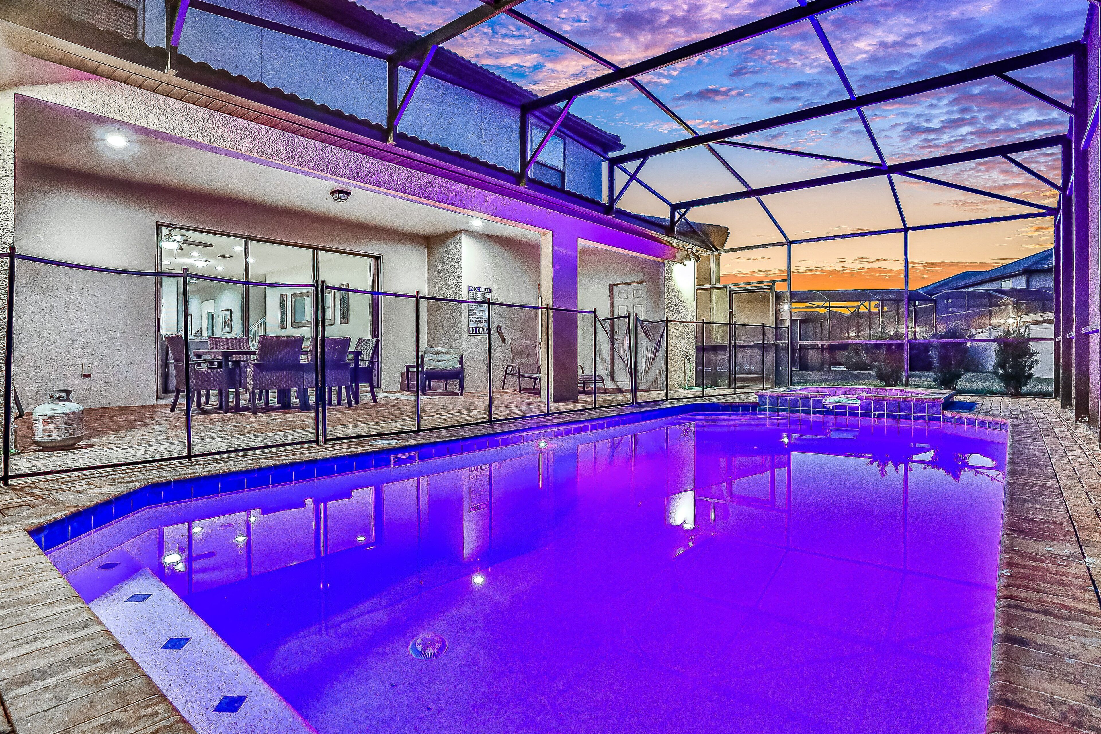 Une piscine entourée de parois en verre, un espace patio aménagé avec du mobilier d’extérieur, et une vue sur le coucher du soleil à travers la structure.