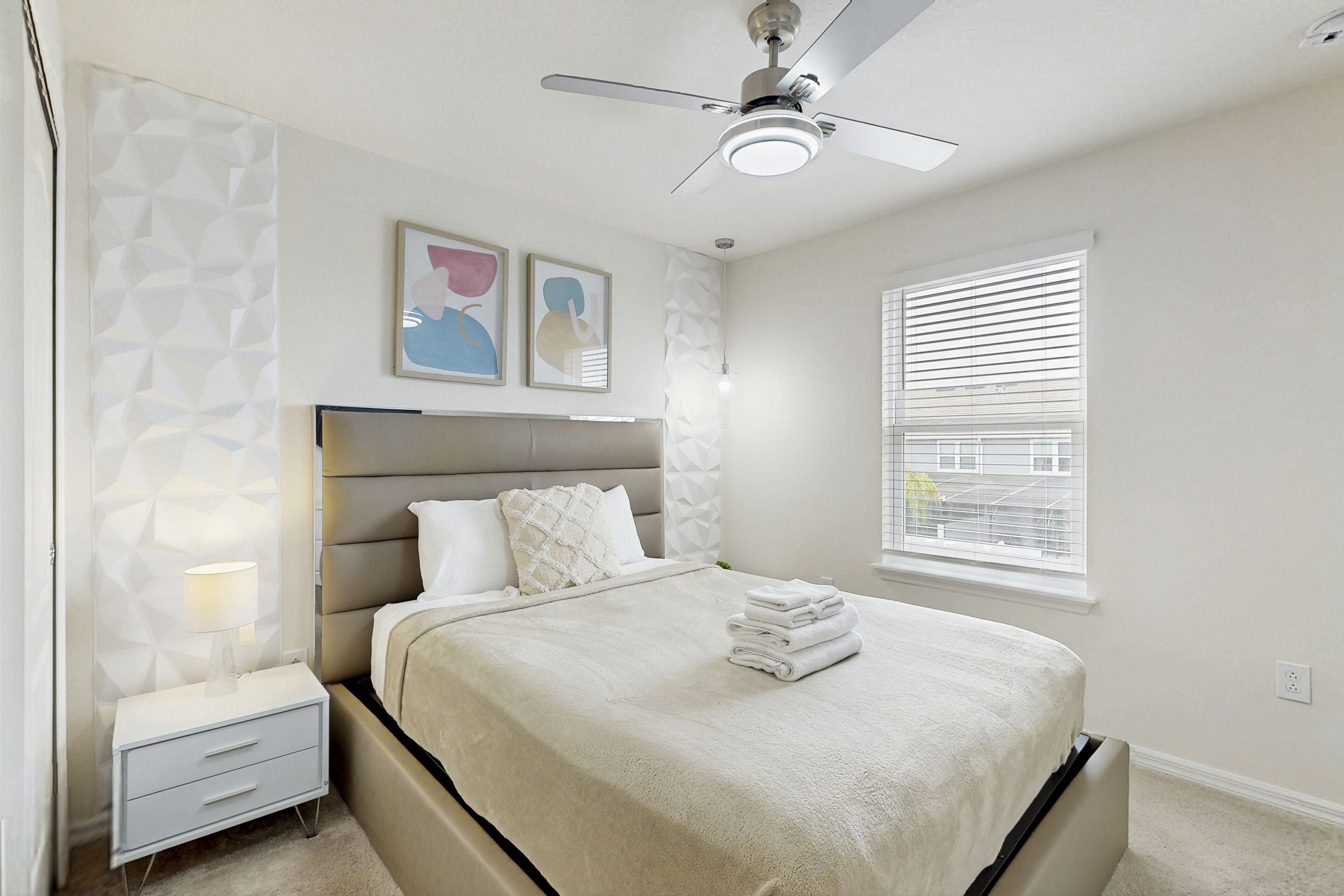 A bedroom with a bed, nightstand, wall art, ceiling fan, and window with blinds.