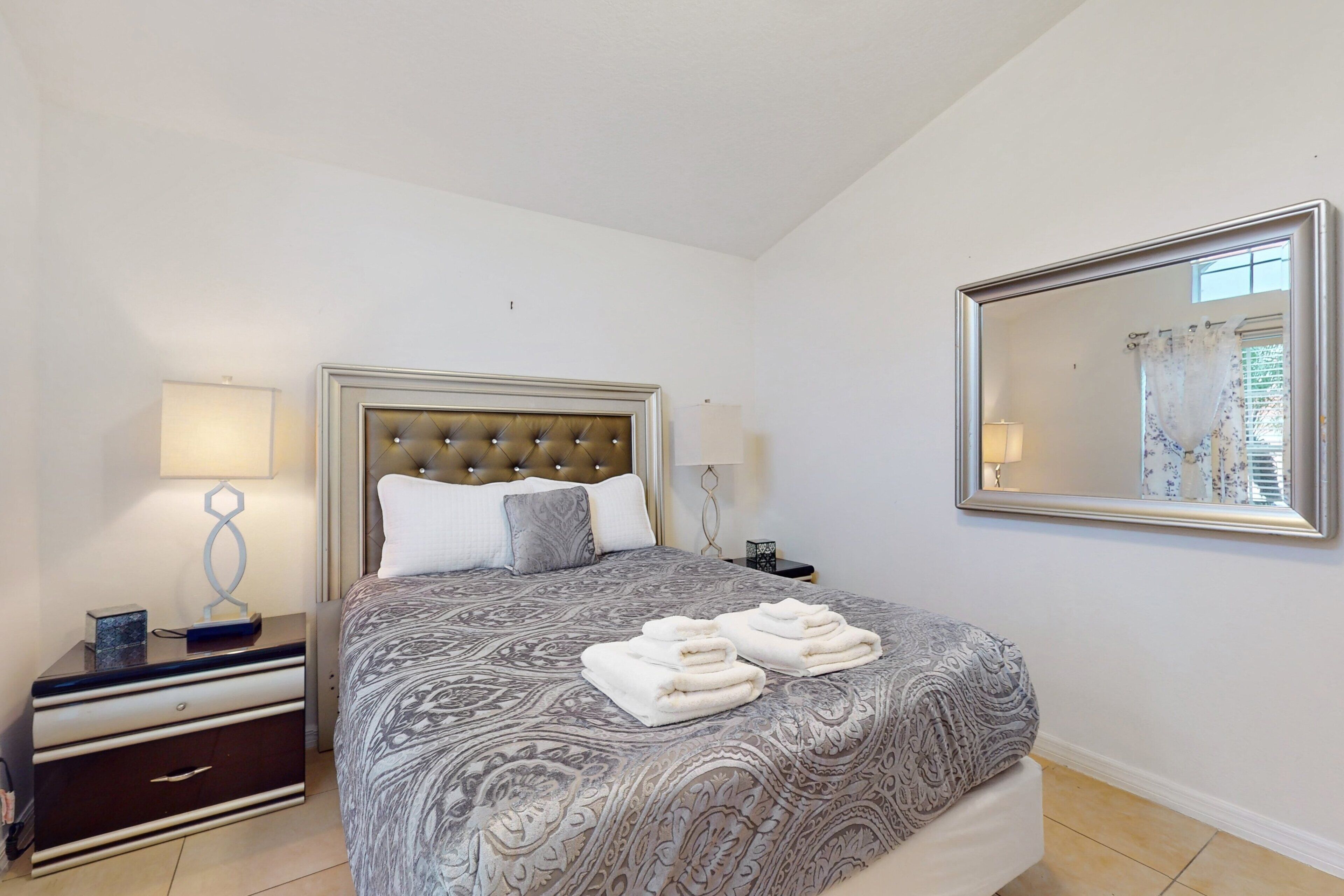 A bedroom with a tufted headboard, a bed with a patterned bedspread, two bedside tables with lamps, and a large mirror on the wall.