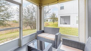 Un espacio de estar al aire libre cubierto con mobiliario de mimbre y una mesa de vidrio, con vista a un patio césped y un edificio.