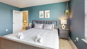 Un dormitorio con cama, mesita de noche, lámpara de pared y dos cuadros enmarcados.