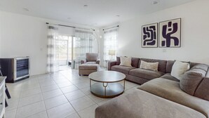 Una sala de estar moderna con un sofá, un reposapiés y una mesa de centro. Hay un televisor y una lámpara sobre una mesita auxiliar.