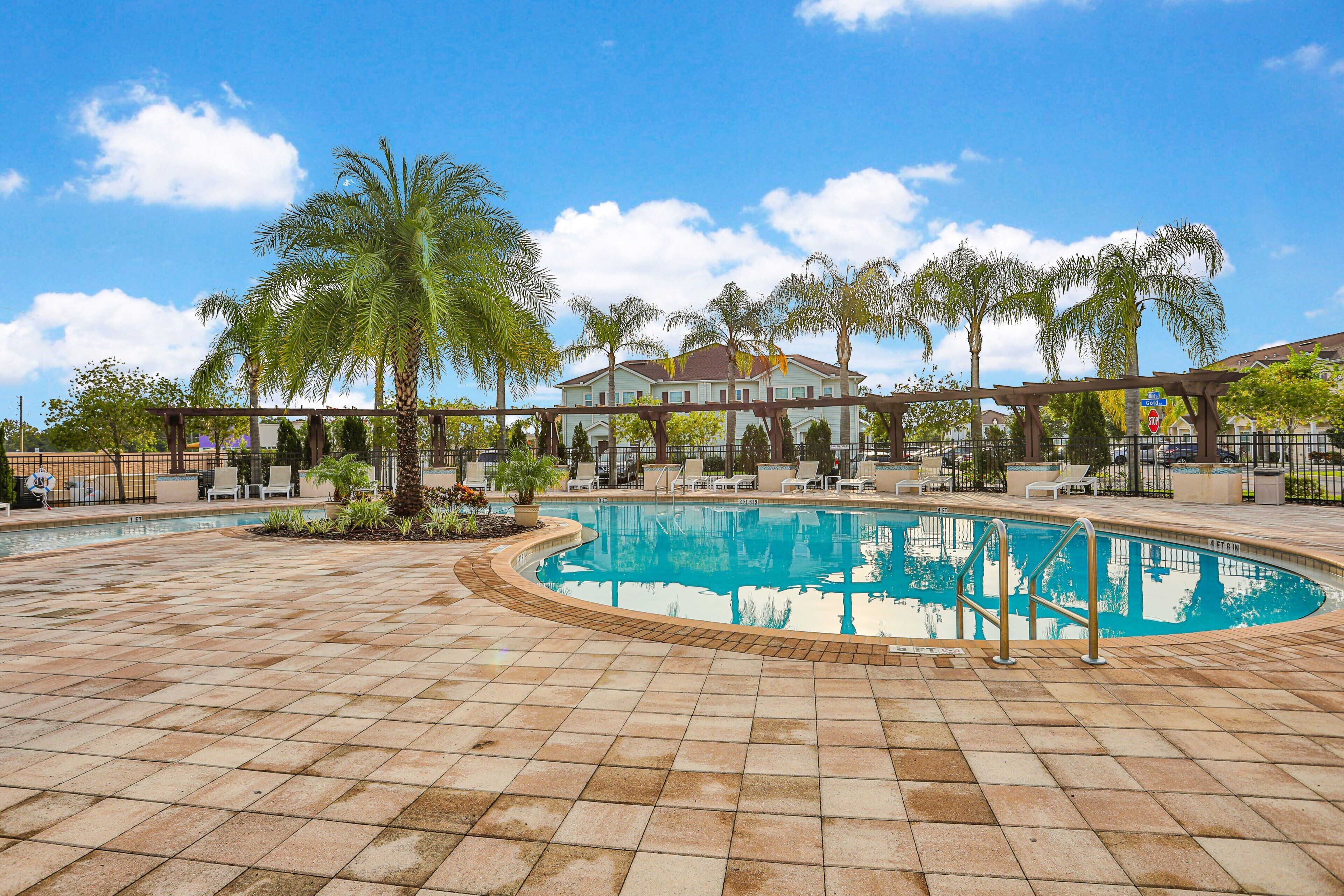 Una piscina con un borde empedrado, palmeras y un edificio al fondo.
