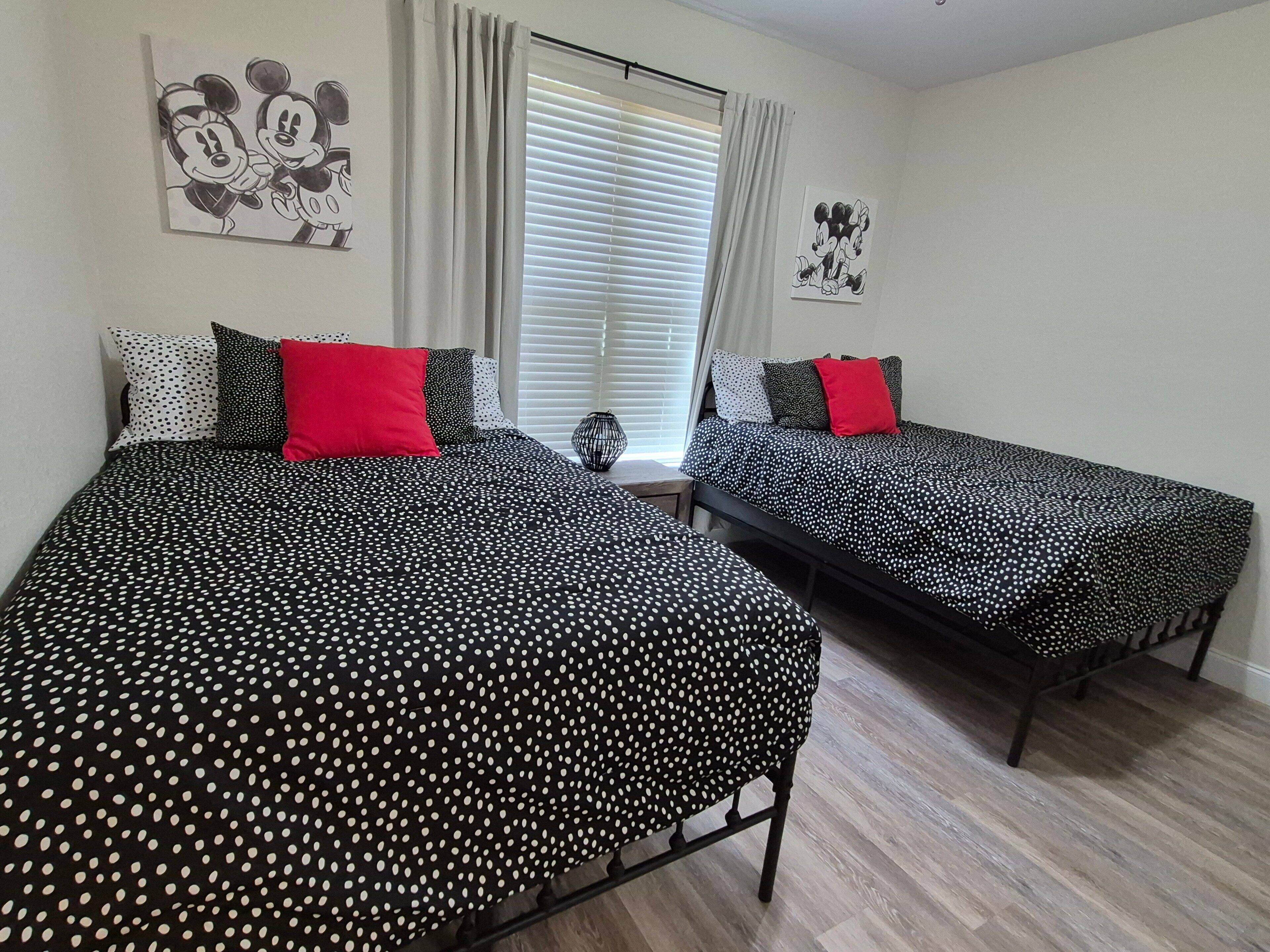Une chambre avec deux lits, chacun agrémenté de draps à pois noirs et blancs et d’oreillers rouges. Au-dessus des lits, on trouve des décorations murales représentant Mickey Mouse.