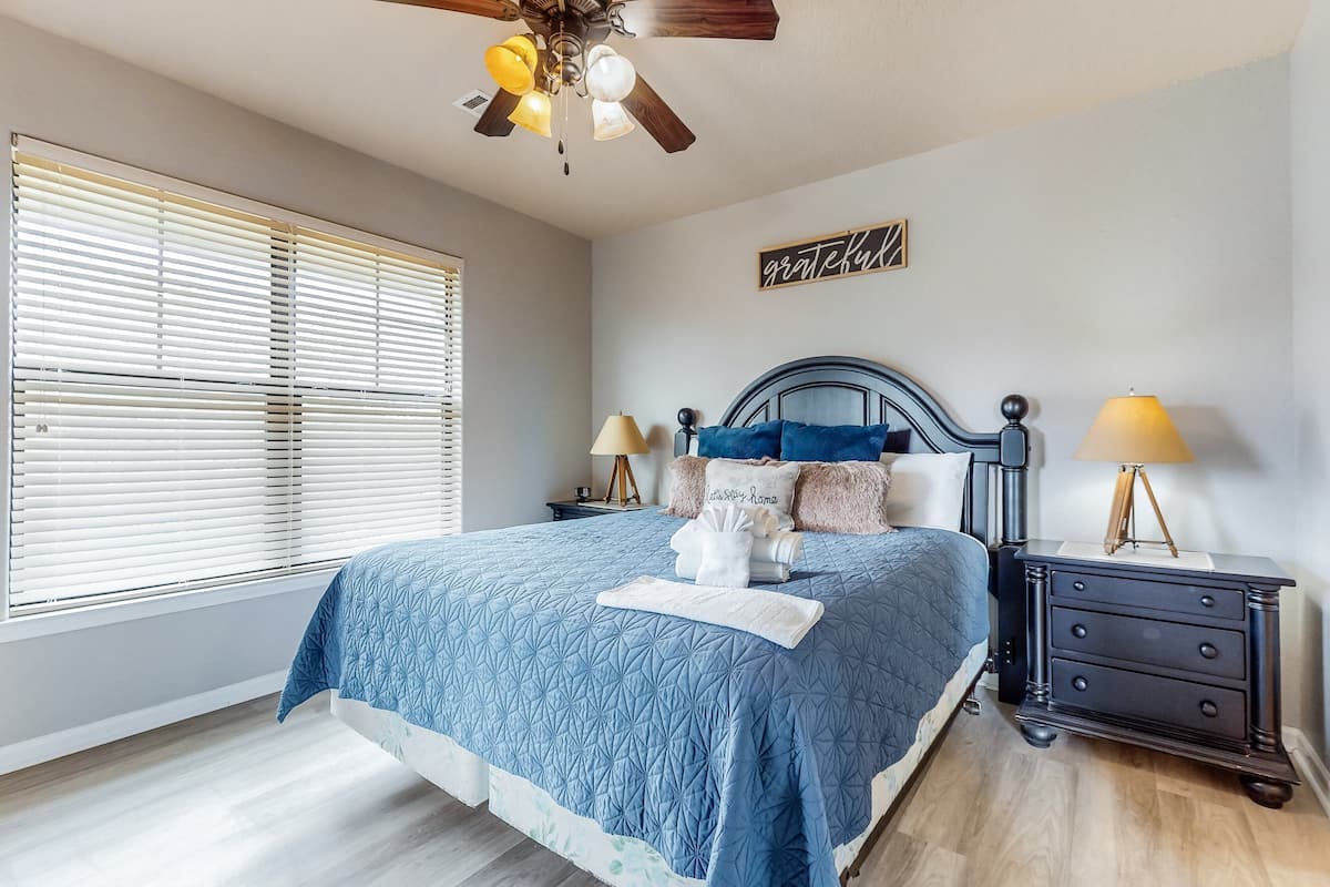 Une chambre à coucher comprenant un lit, une table de chevet, une lampe, un ventilateur de plafond et une fenêtre avec des stores.