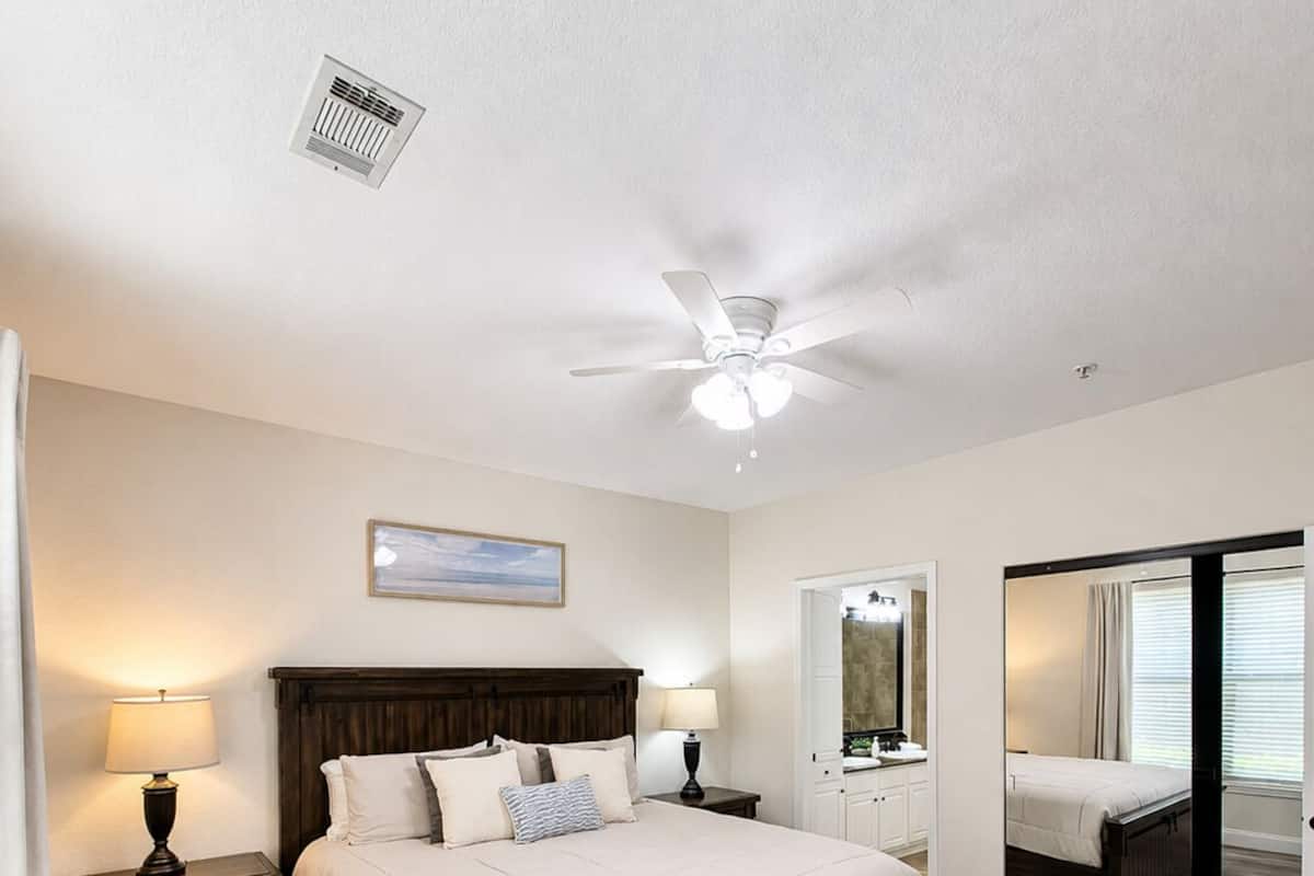Une chambre à coucher comprenant un lit, des tables de chevet, un ventilateur de plafond, un miroir et une porte donnant sur une autre pièce.