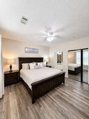 A bedroom with a bed, nightstands, a ceiling fan, a mirror, and a door leading to another room. - Bella Piazza (Davenport)