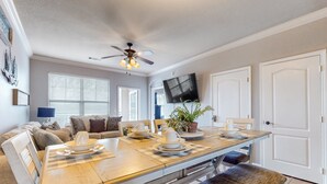 Un coin repas avec une table en bois dressée pour quatre personnes, un ventilateur de plafond, un téléviseur à écran plat fixé au mur et une grande fenêtre équipée de stores.