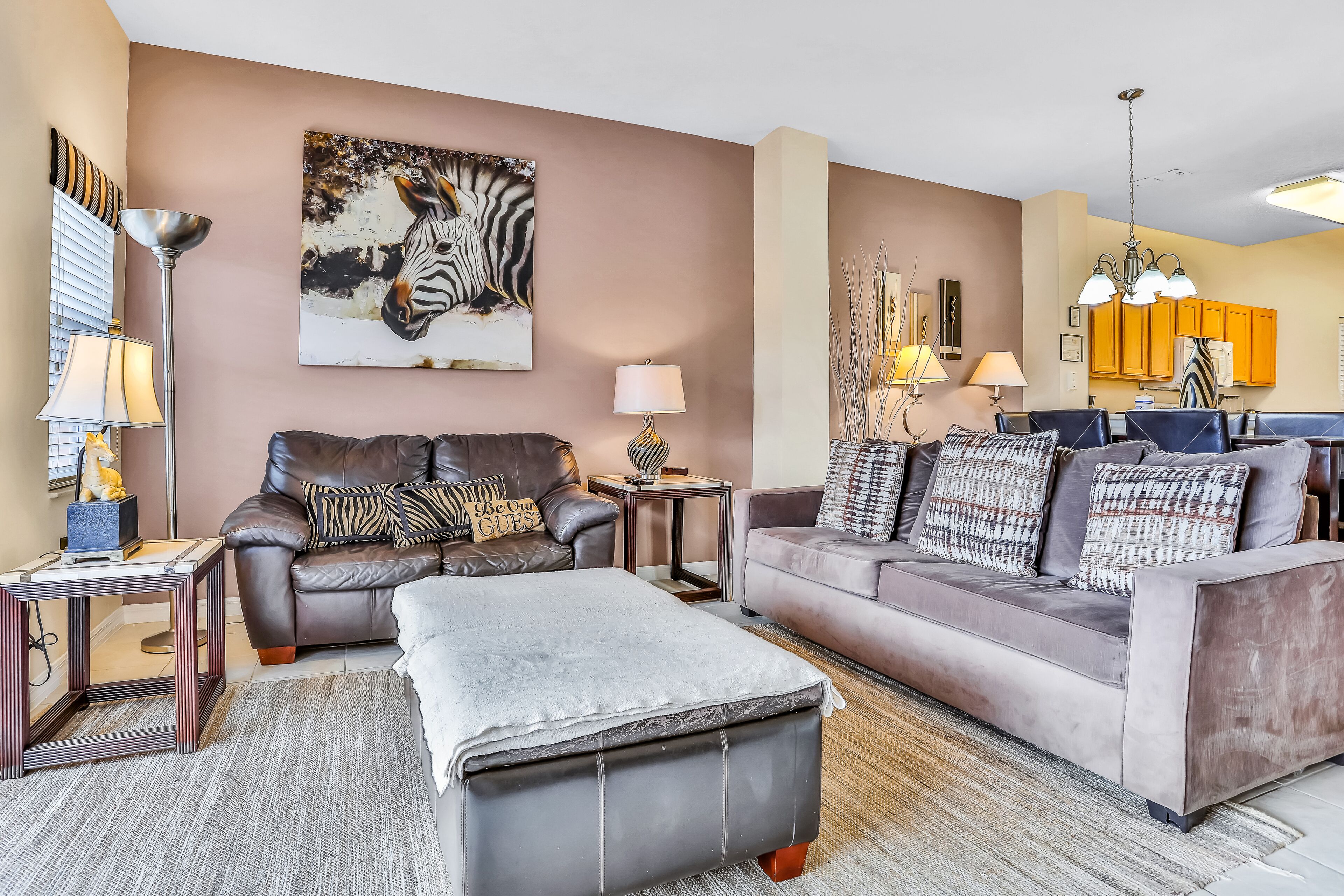 Ein Wohnzimmer mit einer braunen Ledersofa, einem grauen Ottomanen und einem grauen Sofa mit gemusterten Kissen. An der Wand hängt ein Zebra-Gemälde, und es gibt eine Stehlampe sowie eine Tischlampe.