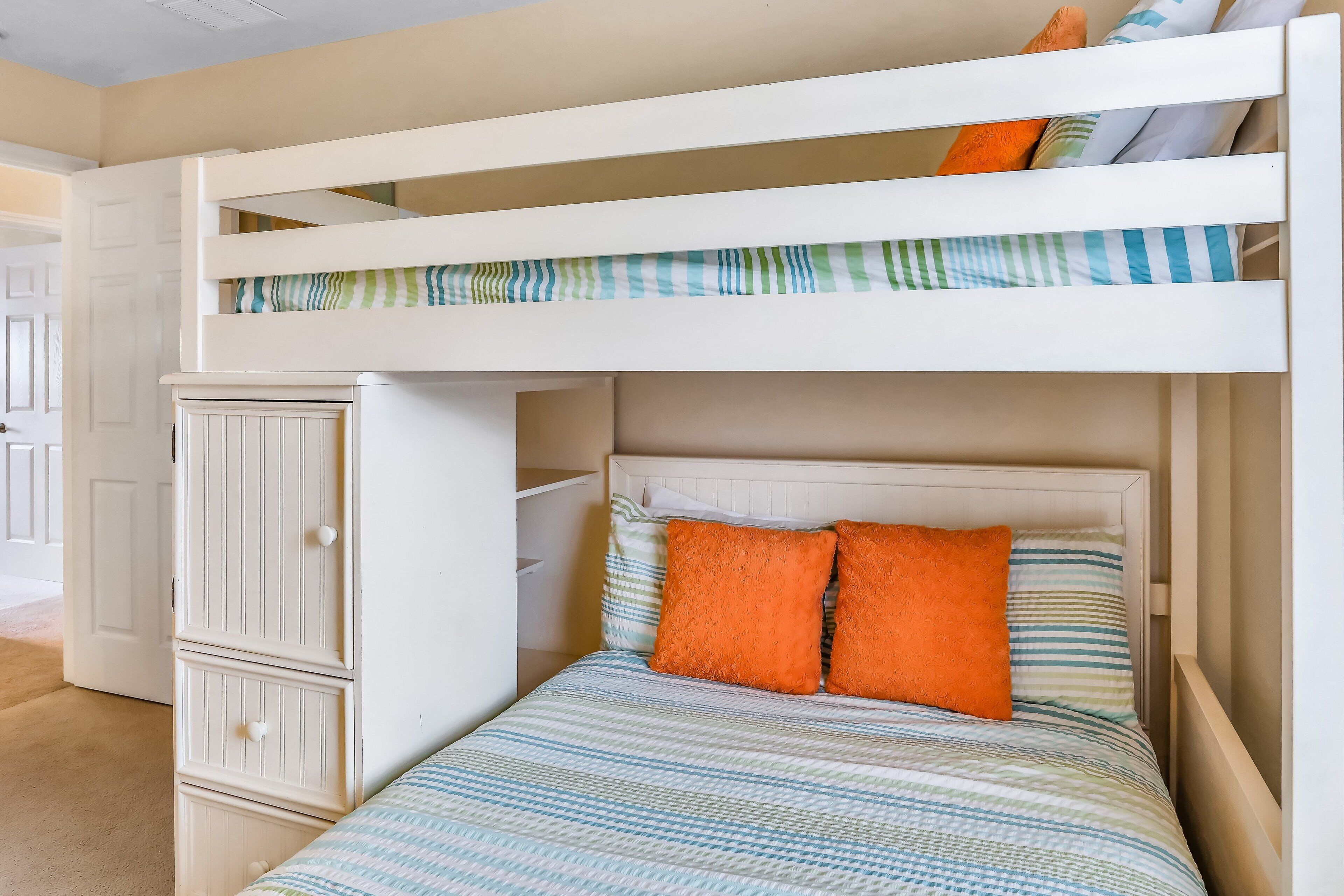 A bunk bed with a lower bed and a storage cabinet, a striped bedspread, and two orange pillows.