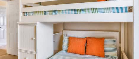 A bunk bed with a lower bed and a storage cabinet, a striped bedspread, and two orange pillows.
