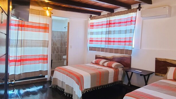Ein Zimmer mit Holzbalkendecke, zwei Betten mit gestreifter Bettwäsche, einem kleinen Tisch und einem Fenster mit Vorhängen.