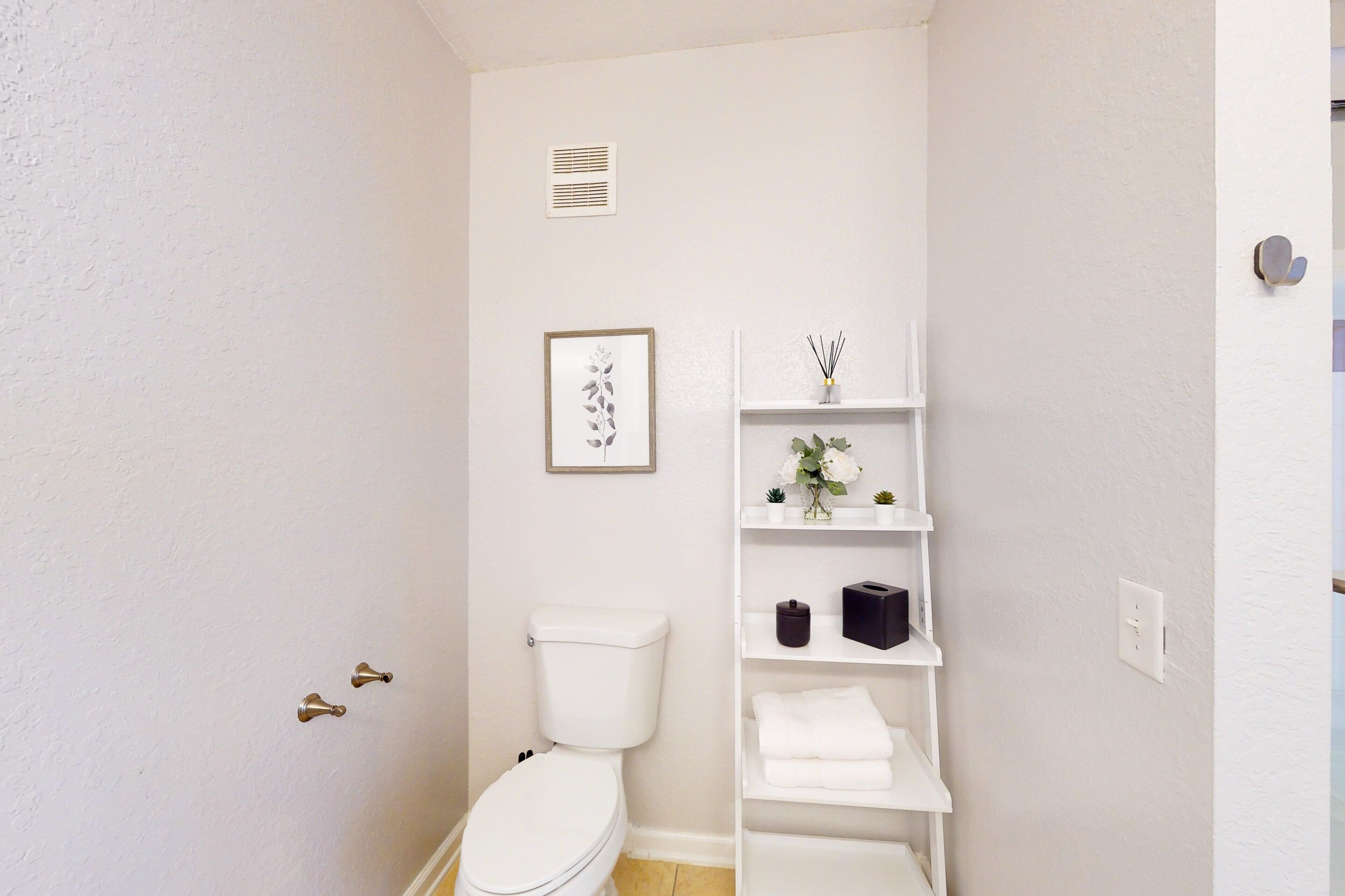 Ein kleines, weißes Badezimmer mit Toilette, einem gerahmten Bild an der Wand und einem weißen Regal mit Vase und kleiner Pflanze.