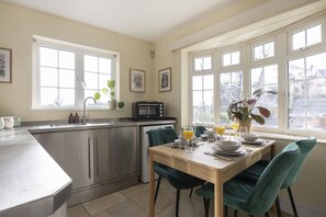 A modern kitchen with stainless steel appliances, a dining table set for four, and large windows offering a view of the outdoors. - Riverside Cottage and Shepherd’s Hut with Hot Tub (Tewkesbury)