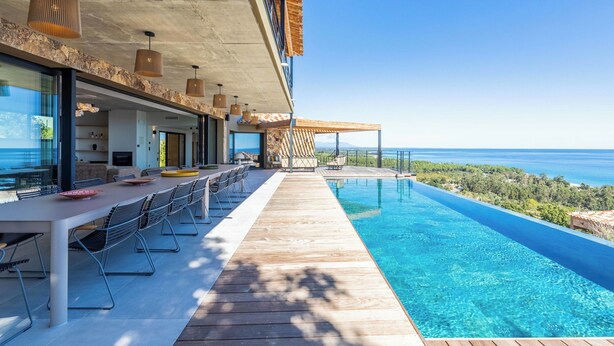 Ein moderner Poolbereich im Freien mit Holzterrasse, einem Esstisch mit Stühlen und Blick aufs Meer.
