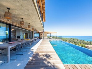 Ein moderner Poolbereich im Freien mit Holzterrasse, einem Esstisch mit Stühlen und Blick aufs Meer.