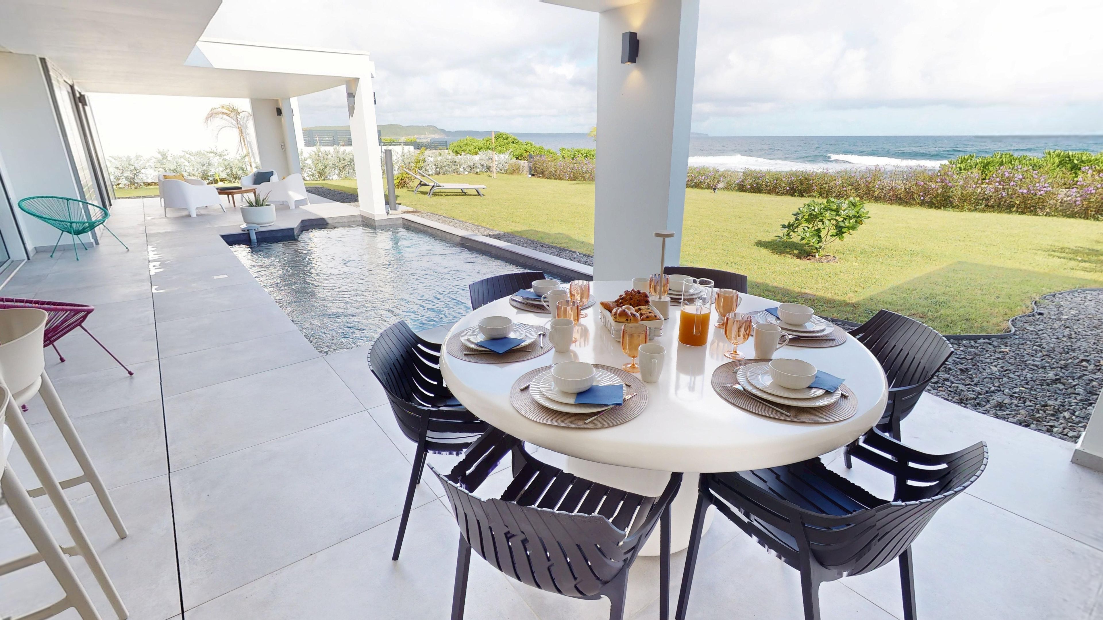 A dining area with a view of the ocean, a pool, and a garden.