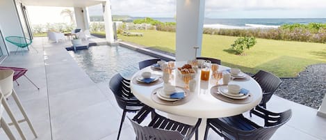 A dining area with a view of the ocean, a pool, and a garden.