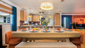 A modern dining area with a large wooden table, orange chairs, and a bench. There is a chandelier above the table, and a neon sign in the background. - Htd Pool, TikiHut, Pickleball, Golf, 7 mi to AMI (Bradenton)