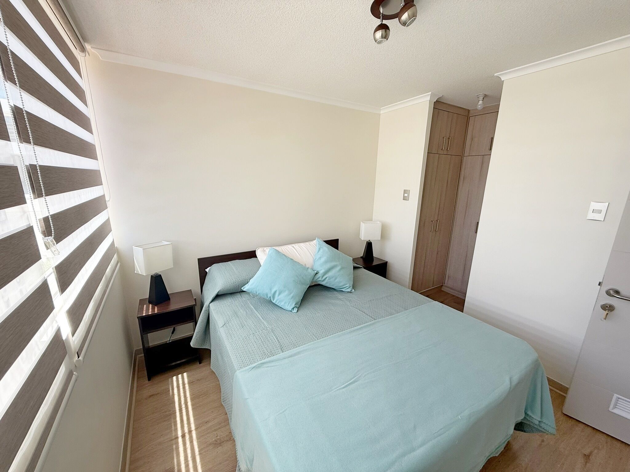 Une chambre à coucher comprenant un lit, des tables de chevet, une armoire et une fenêtre avec des stores.