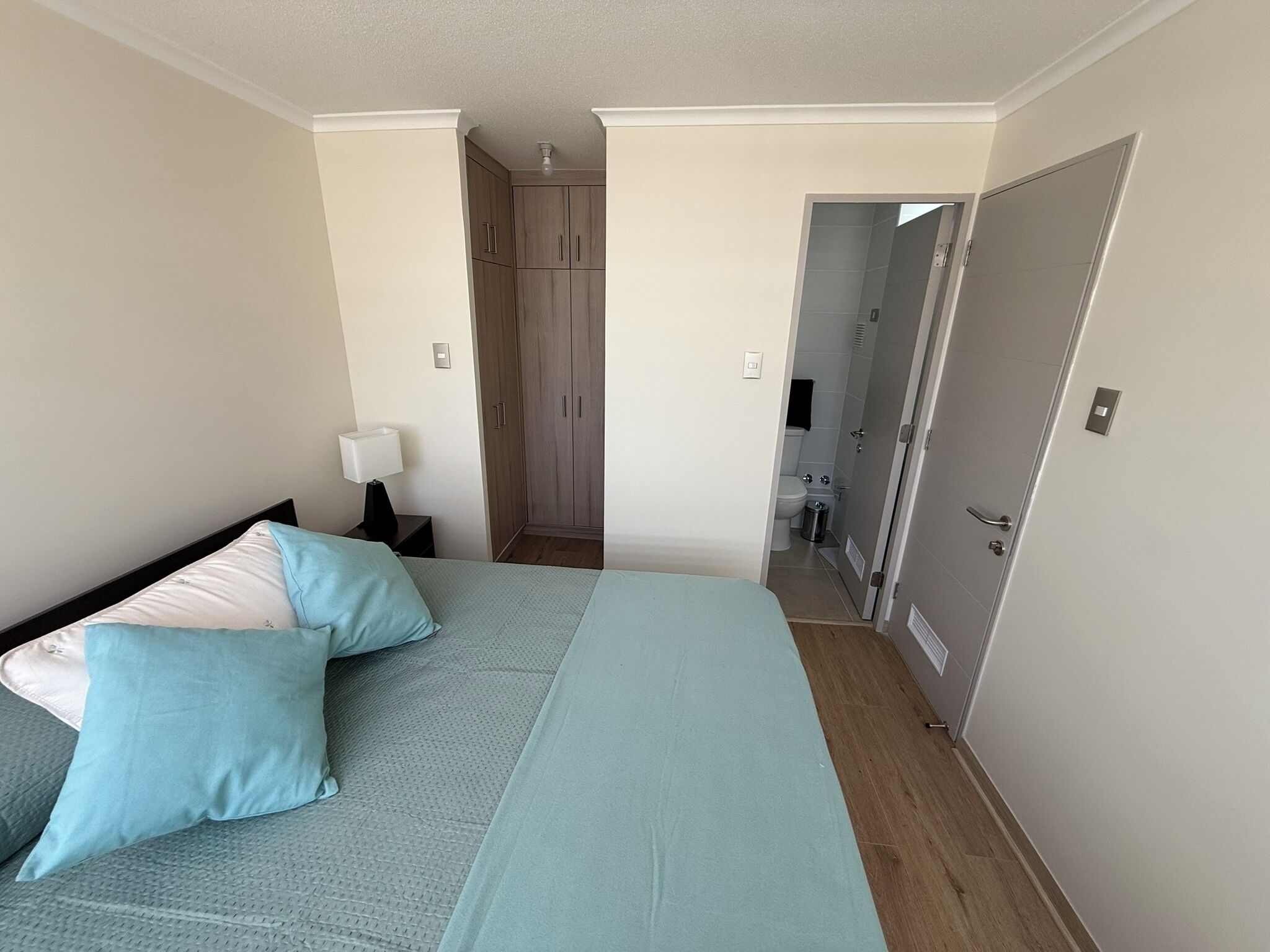 Een slaapkamer met een bed, kussens, een nachtkastje, een kledingkast en een badkamer zichtbaar door een geopende deur.