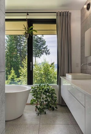 A modern bathroom with a freestanding bathtub, a sink, and a large window with a view of trees. - CEDAR HOME with sauna and cold plunge. 45 min drive from vancouver and Whistler! (Squamish-Lillooet D)