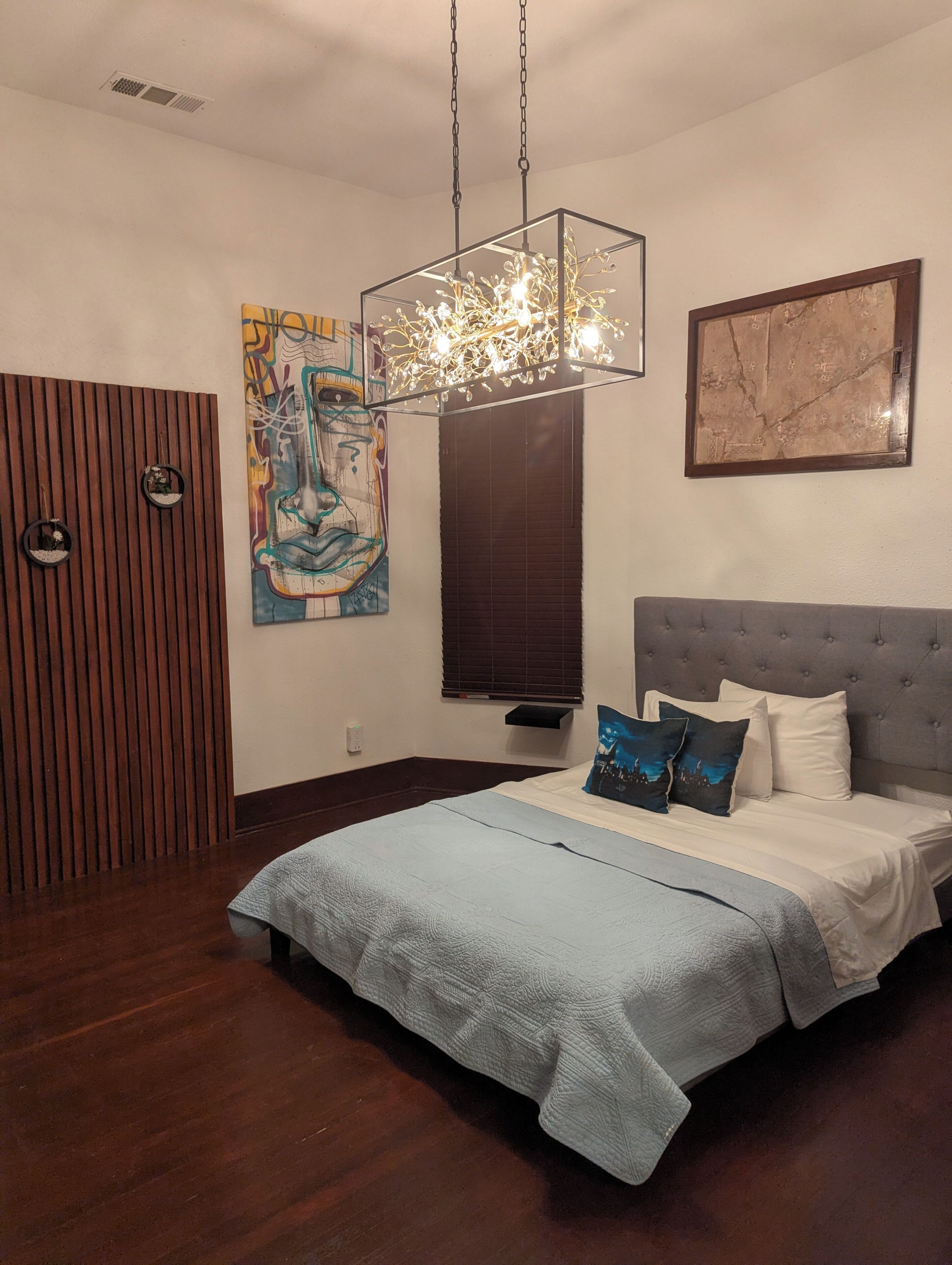 A bedroom with a bed, a chandelier, a wall with a painting, and a window with blinds.