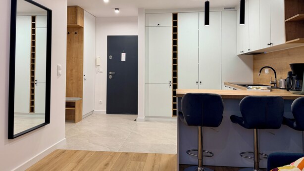 A modern kitchen with a black door, white cabinets, and a bar area with two blue bar stools. - Modern Apartment PKiN Best Location (Warsaw)