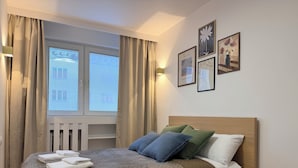 A neatly made bed with a quilted cover, a headboard, and pillows, a window with blinds, and wall art. - Modern Apartment PKiN Best Location (Warsaw)