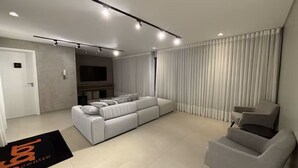 A modern living room with a sectional sofa, a grey armchair, and a flat-screen TV mounted on the wall. - Studio in the city center | Near Passeio Público (Curitiba)