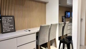 A modern hotel room with a white desk, grey upholstered chairs, a wooden slat wall, and a black sign listing cities. - Studio in the city center | Near Passeio Público (Curitiba)