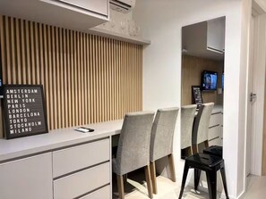 Ein modernes Hotelzimmer mit weißem Schreibtisch, grau gepolsterten Sesseln, einer Wand aus Holzlamellen und einem schwarzen Schild mit aufgelisteten Städten.