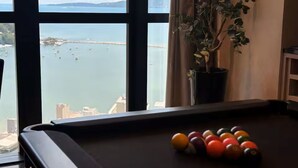 A pool table with balls in front of a window overlooking a body of water and distant buildings. - Apartment with gym, terrace, pool and sea view. (Itapema)