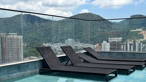 A rooftop pool with lounge chairs, a glass barrier, and a cityscape with mountains in the background. - Apartment with gym, terrace, pool and sea view. (Itapema)