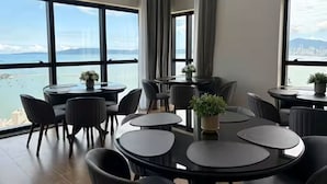 Un comedor moderno con mesas y sillas redondas, ventanas amplias con vistas al mar y al paisaje urbano, y un gran arreglo floral como centro de mesa.