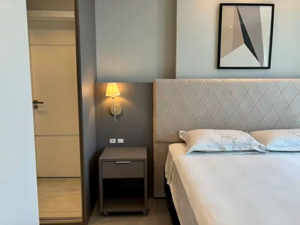 Ein Hotelzimmer mit einem Bett, einem Nachttisch mit Lampe, einem gerahmten Bild an der Wand und einer Tür zu einem anderen Raum.