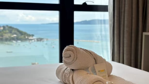 Una cama con toallas enrolladas y una etiqueta, una ventana con vista a una playa y al agua.