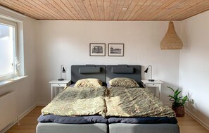 A bedroom with a wooden ceiling, two bedside tables with lamps, a bed with floral bedding, two framed pictures on the wall, and a hanging light fixture.
