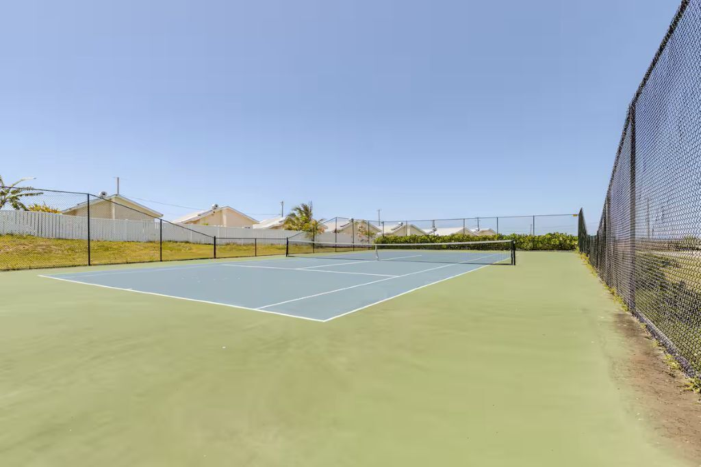Una pista de tenis a l'aire lliure amb xarxa, envoltada per una tanca, i cases al fons.