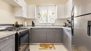 A kitchen with a stove, oven, refrigerator, and a window with a floral curtain. - Sunshine Seashore Escape Jamaica (Lucea)