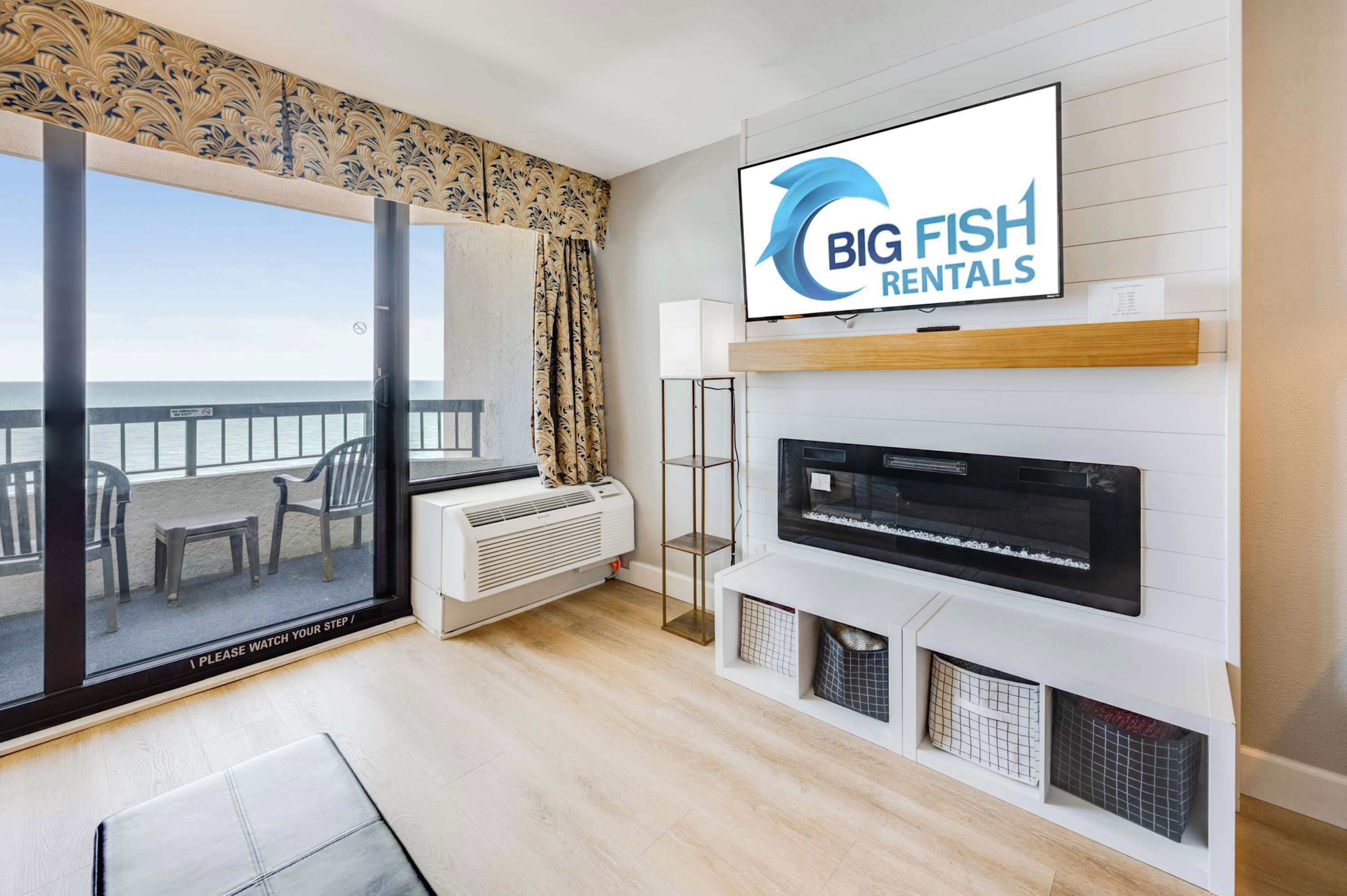 A modern living room with a large TV, a fireplace, a balcony with a view of the sea, and a sign for Big Fish Rentals.