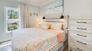 Een slaapkamer met een bed, nachtkastje, dressoir, plafondventilator, raam met gordijnen en een schilderij aan de muur.