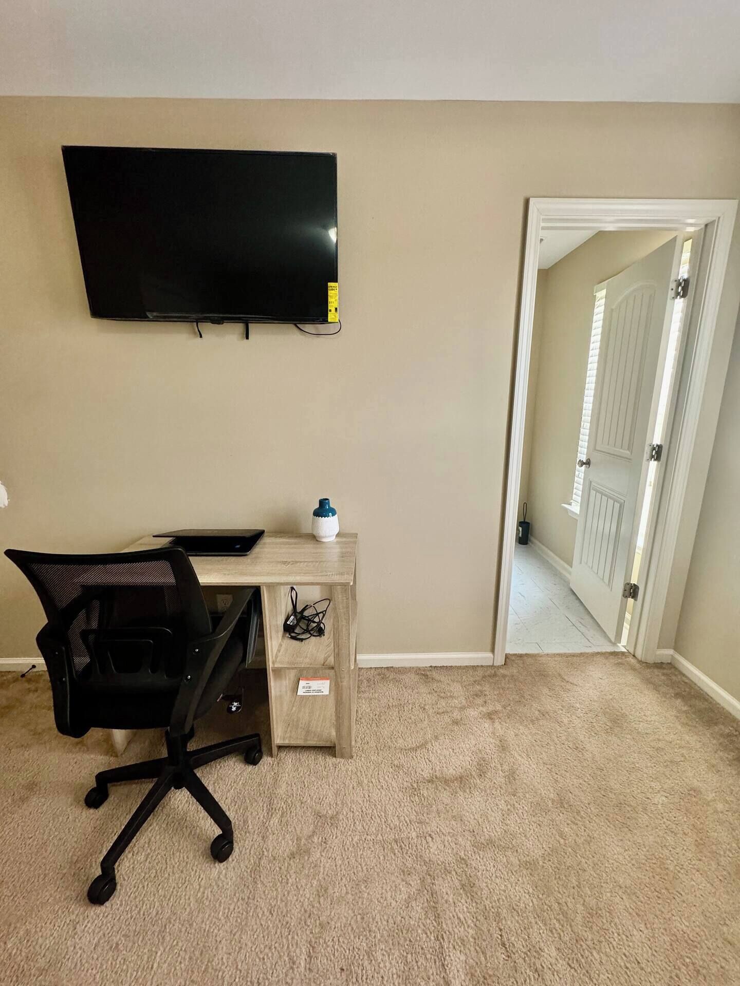 Een kamer met een wandgemonteerde flatscreen-tv, een houten bureau met een stoel, en een deur naar een andere kamer.