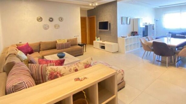 Una sala de estar moderna con sofá, mesa de centro y televisor. Se observa un comedor con sillas y una mesa.