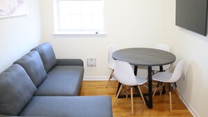 A modern living room with a grey sofa, a round table with chairs, a window with blinds, and a wall-mounted TV. - Beautiful 1Bed 1 Bath w/ Parking (Belleville)