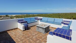 A rooftop terrace with a blue patterned sofa set, a stone fire pit, and a view of the ocean.