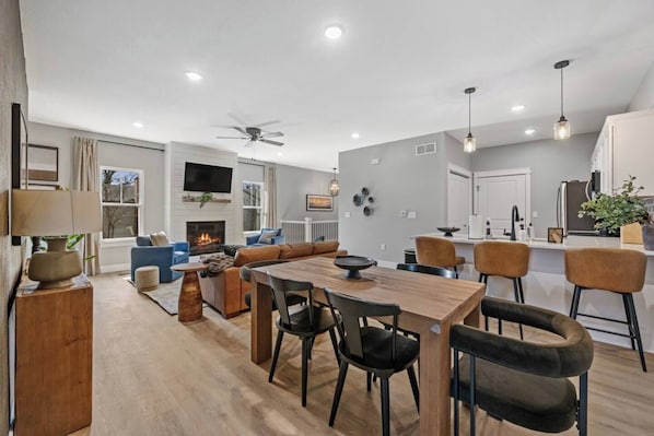 A modern living room with a fireplace, a dining area with a wooden table and chairs, a kitchen with white cabinets, and a ceiling fan.